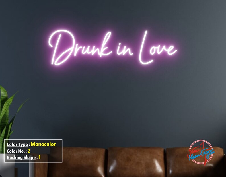May include: Pink neon sign with the text "Drunk in Love" on a dark gray wall. The sign is mounted above a brown leather couch.