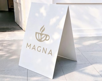 May include: A white A-frame sidewalk sign featuring a beige graphic of a coffee cup with steam rising from it. Below the graphic, the word MAGNA is printed in large, bold, beige capital letters. The sign has a smooth, matte finish.