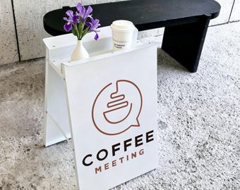 May include: A white A-frame sign featuring a brown circular logo and the text COFFEE MEETING. A white vase with purple iris flowers and a white disposable coffee cup with a lid sit on the top shelf of the sign, which is positioned on a textured floor.