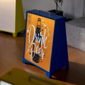 May include: A-frame sign with a blue frame and a handle. The sign features an orange background with a black beer bottle illustration and the words "Drink Beer" in white script. White paint splatters add a decorative touch.