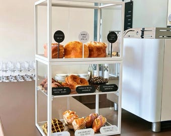 May include: A two-tiered white metal and glass display case containing various baked goods. The top shelf holds three round pastries with circular labels. The middle and bottom shelves feature assorted croissants, pretzels, and waffles with small black labels.