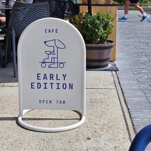 Op de afbeelding: Een wit, boogvormig cafébord met een blauw hondenlogo en tekst. Het bord leest "CAFE" bovenaan, met "EARLY EDITION" en "OPEN 7 AM" eronder. Het bord staat op een stoep.