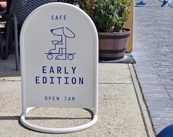 May include: A white, arched cafe sign with a blue dog logo and text. The sign reads "CAFE" at the top, with "EARLY EDITION" and "OPEN 7 AM" below. The sign is on a sidewalk.