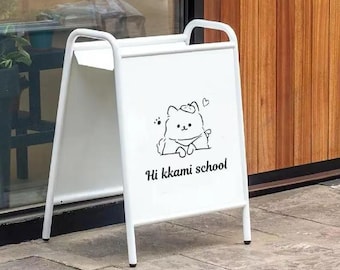 May include: A white A-frame sign with a cartoon bear illustration and the text "Hi kkami school." The sign has a simple, clean design with a metal frame and is placed on a concrete surface.