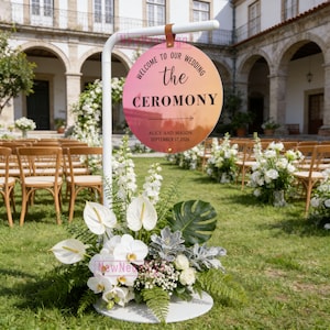 May include: A wedding ceremony sign with a circular design, featuring text that reads "Welcome to our Wedding the Ceremony." The sign is supported by a white pole and adorned with floral arrangements. The sign also includes the names Alice and Mason and the date September 17, 2026.