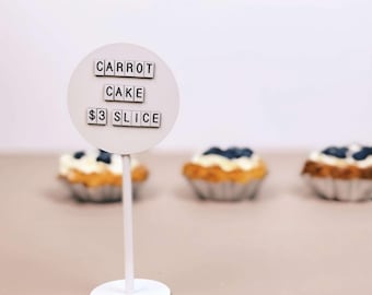 May include: A circular white sign on a thin stand displaying the text CARROT CAKE $3 SLICE in black letters. The sign is positioned in front of three small baked goods topped with white frosting and blueberries, arranged in a horizontal row.