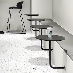 Puede incluir: Un interior moderno con un taburete de bar negro y varias mesas pequeñas, redondas y negras unidas a un banco blanco. Una taza de café desechable está sobre una mesa. El suelo es blanco con patrones moteados.