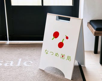 May include: A white A-frame sidewalk sign featuring a graphic of three red cherries with green stems. Below the cherries, the Japanese text "なつめ薬局" is printed in green. The sign has a cutout handle at the top and a curved cutout at the base.