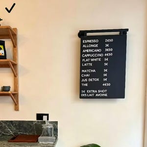 Puede incluir: Una pizarra negra con un menú de café y té en texto blanco. El menú incluye espresso, latte y matcha. La pizarra está colgada en una pared de color claro.