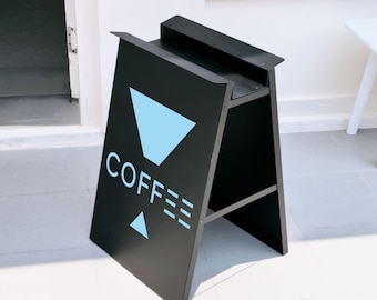 May include: A black A-frame sidewalk sign featuring a light blue graphic of a coffee dripper above the word COFFEE in light blue capital letters, with a small light blue triangle below the text, constructed from metal with a matte finish.