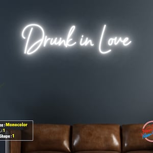 May include: White neon sign with the text "Drunk in Love" on a dark gray wall. The sign is mounted above a brown leather couch.