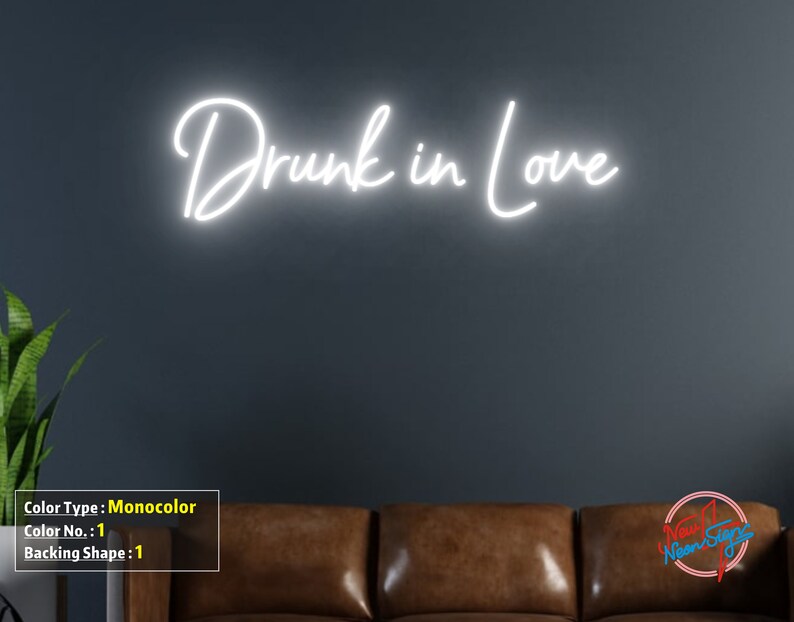 May include: White neon sign with the text "Drunk in Love" on a dark gray wall. The sign is mounted above a brown leather couch.