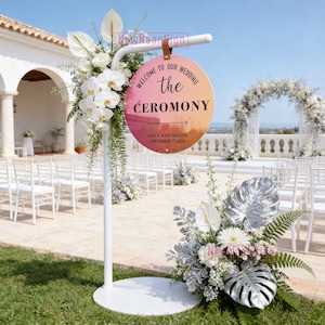 Może przedstawiać: Biały metalowy znak weselny z okrągłym znakiem z gradientem od różu do pomarańczu. Znak głosi "Welcome to our Wedding the Ceremony" z imionami i datą. Ozdobiony białymi kwiatami i zielenią.