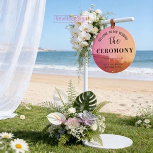 Puede incluir: Un letrero de ceremonia de boda con un círculo degradado rosa y naranja que dice "Bienvenida a nuestra boda la ceremonia" con los nombres de Alice y Mason y la fecha 17 de septiembre de 2026. El letrero está decorado con flores y vegetación.