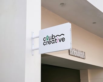 May include: A white rectangular double-sided projecting sign mounted on a white wall. The sign features the black text club creative with a green letter c, a pink letter i, and a black wavy line above the word creative. The sign is attached by two white metal arms.
