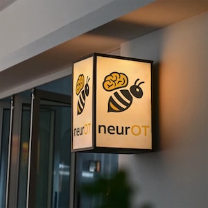 Puede incluir: Un letrero rectangular iluminado con un marco negro. El letrero presenta un gráfico de abeja amarilla y negra con un cerebro encima. El texto "neurOT" está debajo de la abeja en amarillo. El letrero está montado en una pared.