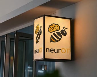 May include: A rectangular, illuminated sign with a black frame. The sign features a yellow and black bee graphic with a brain on top. The text "neurOT" is below the bee in yellow. The sign is mounted on a wall.