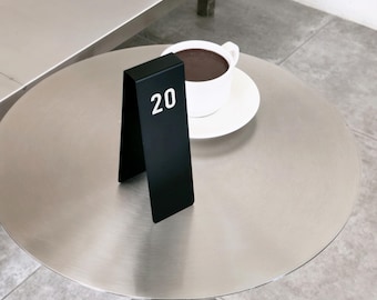 May include: A matte black metal table number sign featuring the white printed number 20 in a bold sans-serif font. The sign is folded into an inverted V-shape and stands upright next to a white ceramic cup filled with dark coffee on a saucer.
