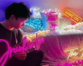 May include: A person holding a pink neon sign that says "Drunk in Love". Other neon signs in the background include "Game On", "The Custom's LED Sign", and "Till Death". The signs are in various colors.