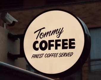 May include: A round, illuminated sign for "Tommy Coffee" with black text on a cream background. The sign reads "Tommy COFFEE FINEST COFFEE SERVED" in a stylized font. The sign is mounted on a dark metal bracket.