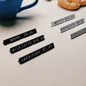 Op de afbeelding: Een verzameling L-vormige metalen bordjes voor eten en drinken in zwart en wit. De zwarte bordjes tonen MOCHA $8.00, MILK LATTE $12.00 en AMERICANO $7.00. De witte bordjes tonen MUFFIN $12.00, CAFE LATTE $9.00 en CAPPUCCINO.