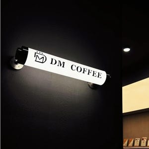 May include: A white, cylindrical light fixture with the text "DM COFFEE" in black lettering is mounted on a dark wall. The fixture is illuminated, and the logo is visible. The fixture is held in place by two silver metal brackets.