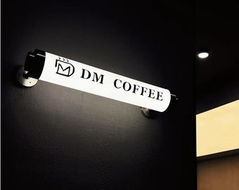 May include: A white, cylindrical light fixture with the text "DM COFFEE" in black lettering is mounted on a dark wall. The fixture is illuminated, and the logo of a stylized "M" is also visible. The fixture is attached to the wall with two silver brackets.