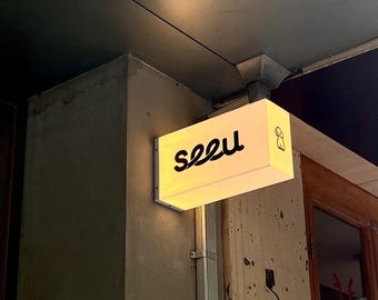 May include: Illuminated rectangular sign with the word "seeu" in black script. A small dog illustration is on the right side of the sign. The sign is mounted on a textured concrete wall, near a wooden door.