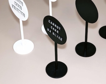 May include: A collection of circular tabletop signs on thin vertical stands with round bases. The signs are available in solid white or black. Each sign features horizontal tracks holding individual letter tiles that spell out CARROT CAKE and the price per slice.