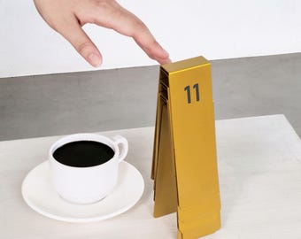 May include: A white ceramic coffee cup and saucer filled with black coffee next to a gold-colored metal table number stand. The stand is folded into a triangular shape and features the black printed number 11 on its front-facing panel.