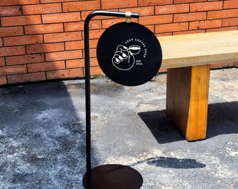 May include: A black metal floor sign featuring a circular hanging panel with a white graphic of a hand holding a coffee cup. The text reads YOUR COFFEE SHOP and estd 2025. The sign is supported by a vertical pole and a round base stand.