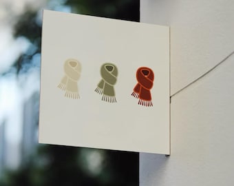 May include: A white square sign displays three scarf illustrations in cream, olive green, and rust-red. Each scarf has a fringed bottom. The sign is mounted on a white wall, with a blurred outdoor background visible.