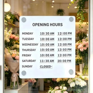 May include: A white rectangular sign with rounded corners and four grey mounting bolts displays the text OPENING HOURS followed by a weekly schedule. Monday through Friday hours are 10:30 AM to 12:00 PM and 10:00 AM to 12:00 PM, Saturday 11:30 AM to 20:00 PM, and Sunday CLOSED.
