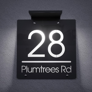 Puede incluir: Un letrero de dirección negro con el número 28 en letras blancas brillantes e iluminadas. El texto "Plumtrees Rd" está debajo del número. El letrero está montado en una pared gris texturizada.