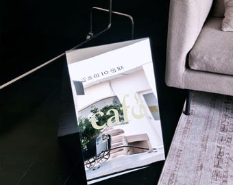 May include: A white A-frame sidewalk sign featuring a printed image of a cafe interior with a vintage-style black carriage, stacked boxes, and the word café in large light-colored lettering. The sign is positioned on a dark floor near a sofa.