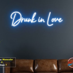 May include: White neon sign with the text "Drunk in Love" on a dark gray wall. The sign is mounted above a brown leather couch.