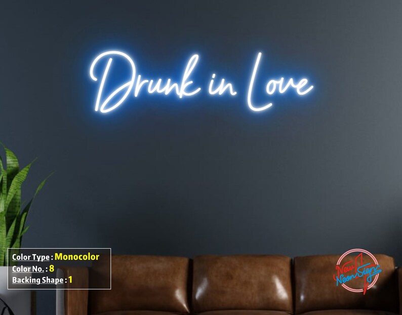 May include: White neon sign with the text "Drunk in Love" on a dark gray wall. The sign is mounted above a brown leather couch.