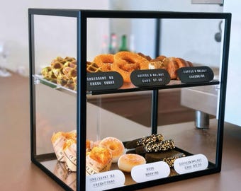 May include: A two-tier black metal and glass display case containing various baked goods. The top shelf holds waffles and pretzels with black labels reading "CROISSANT $3 FRESH TODAY" and "COFFEE & WALNUT CAKE $7.00". The bottom shelf holds pastries.