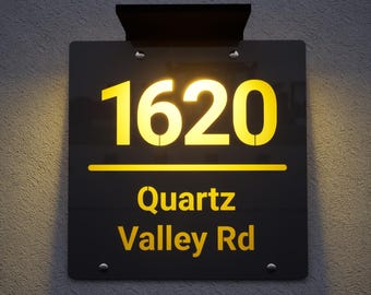 May include: A black illuminated address sign with the number 1620 in a bright yellow font. The text "Quartz Valley Rd" is below the number. The sign is mounted on a textured gray wall.