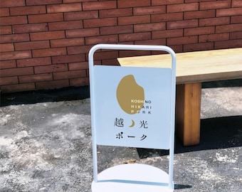 May include: A white metal A-frame sign with a circular base and a rectangular panel. The panel features a gold organic shape, the text KOSHINO HIKARI PORK, and the Japanese characters 越光ポーク with a small crescent moon symbol between the characters.