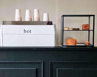 May include: A white espresso machine with the word hot printed on the front, topped with four stacked paper cups. Beside it sits a black metal display case with two glass shelves containing various baked goods, including bread and pastries.