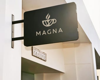 May include: A rectangular black wall-mounted sign featuring a white coffee cup icon and the text MAGNA in capital letters. Below the main sign, a smaller rectangular wooden plaque is mounted to the wall with the word GRATEFUL printed on it.