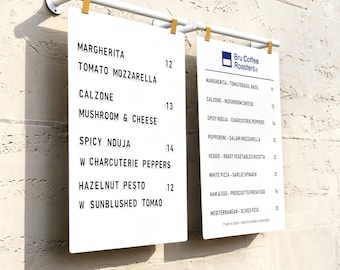 May include: Two white rectangular menu boards suspended from a white horizontal bar with gold clips. The boards feature black text listing food items like Margherita, Calzone, Spicy Nduja, and Hazelnut Pesto, along with prices and Bru Coffee Roasters branding.