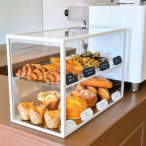 May include: A rectangular white metal and glass display case containing two shelves of assorted baked goods. The top shelf holds croissants, waffles, and dark pastries. The bottom shelf features muffins in paper liners and various golden-brown breads.