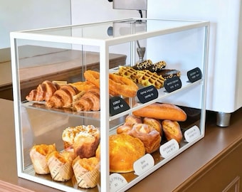 May include: A rectangular white metal and glass display case containing two shelves of assorted baked goods. The top shelf holds croissants, waffles, and dark pastries. The bottom shelf features muffins in paper liners and various golden-brown breads.