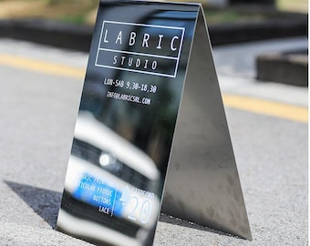 May include: A metallic A-frame sign with the text "LABRIC STUDIO" in white. The sign also displays business hours and an email address. Additional text includes "BRIC PRINTING, TICULAR FABRIC, BOTTONS, LACE, CITOFONO, 20". The sign is placed on a concrete surface.