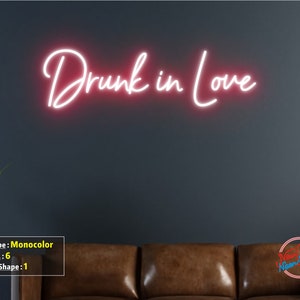 May include: Pink neon sign with the text "Drunk in Love" on a dark gray wall above a brown leather couch.