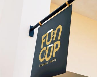 May include: A rectangular black hanging sign mounted on a black metal arm with gold accents. The sign features the gold text FUN CUP with stylized cup graphics integrated into the letters, and the words CERAMIC MUGS printed in smaller gold font.