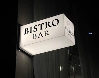 May include: A rectangular, illuminated sign for a bistro bar. The sign is white with the words "BISTRO BAR" in bold, black capital letters. A smaller logo with a wine glass and the words "Bistro Bar" is on the side. The sign is mounted on a dark wall.