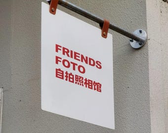 May include: A white rectangular sign with the words "FRIENDS FOTO 自拍照相馆" in red lettering. The sign is suspended from a metal bar with brown leather straps and mounted on a gray wall.